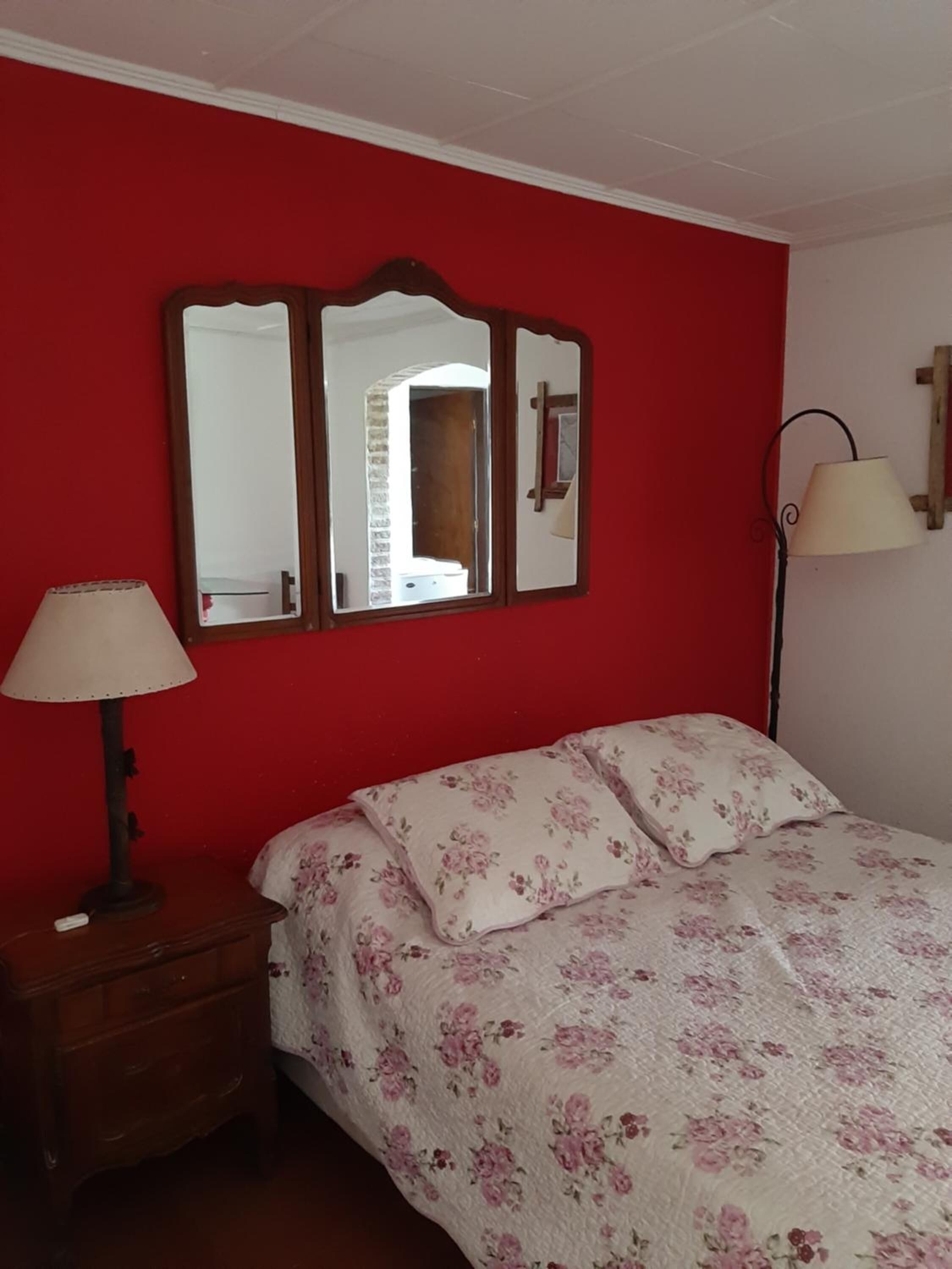 Una habitación con pared roja, una cama con ropa de cama floral, una mesita de noche con una lámpara y un espejo sobre la cama.