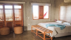 Un dormitorio con una cama, un banco, dos taburetes redondos, un ventanal con contraventanas de madera y suelo de hormigón.