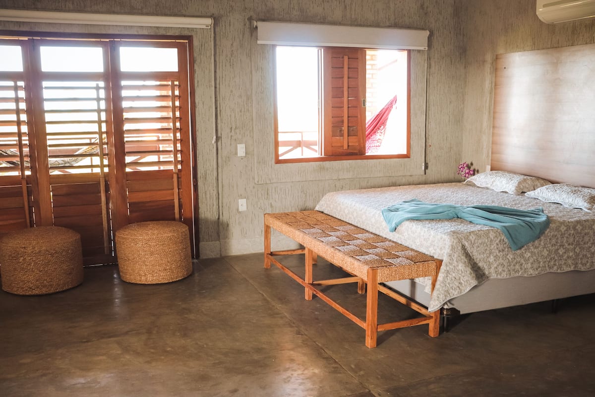 A bedroom with a bed, a bench, two round stools, a window with wooden shutters, and a concrete floor.