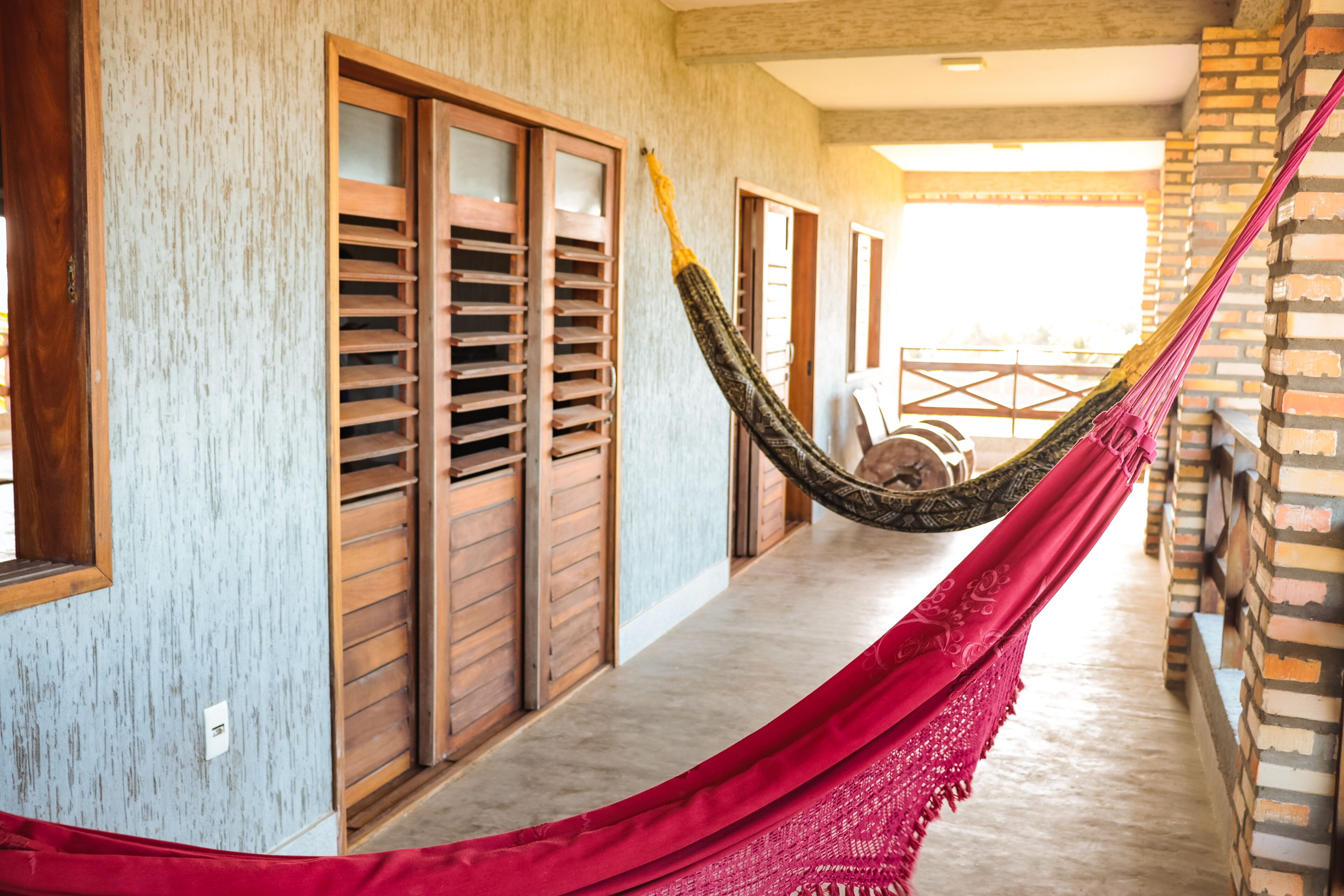 A hammock strung between wooden shutters in a corridor with a brick wall and a view of the outside.