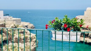 Ein Balkon mit einem weißen Blumentopf, der rote Blumen enthält, mit Blick auf eine felsige Küstenlinie und das Meer.