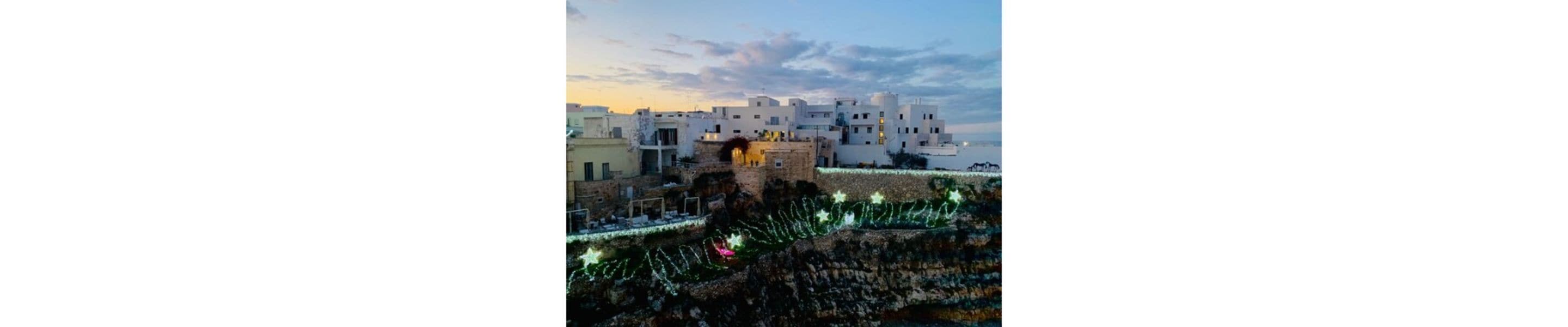 A coastal town with white buildings, a stone wall, and festive lights.