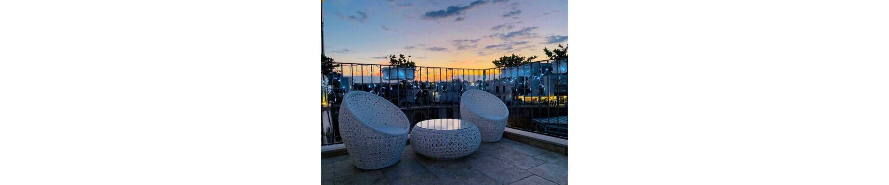 A terrace with three round, perforated chairs and a table, overlooking a cityscape at dusk.