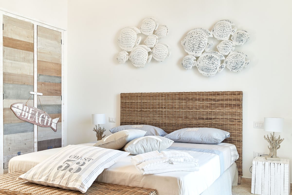 Une chambre à coucher avec un lit, des oreillers, une œuvre d’art murale, un panneau indiquant « Plage » et une table de chevet avec une lampe.