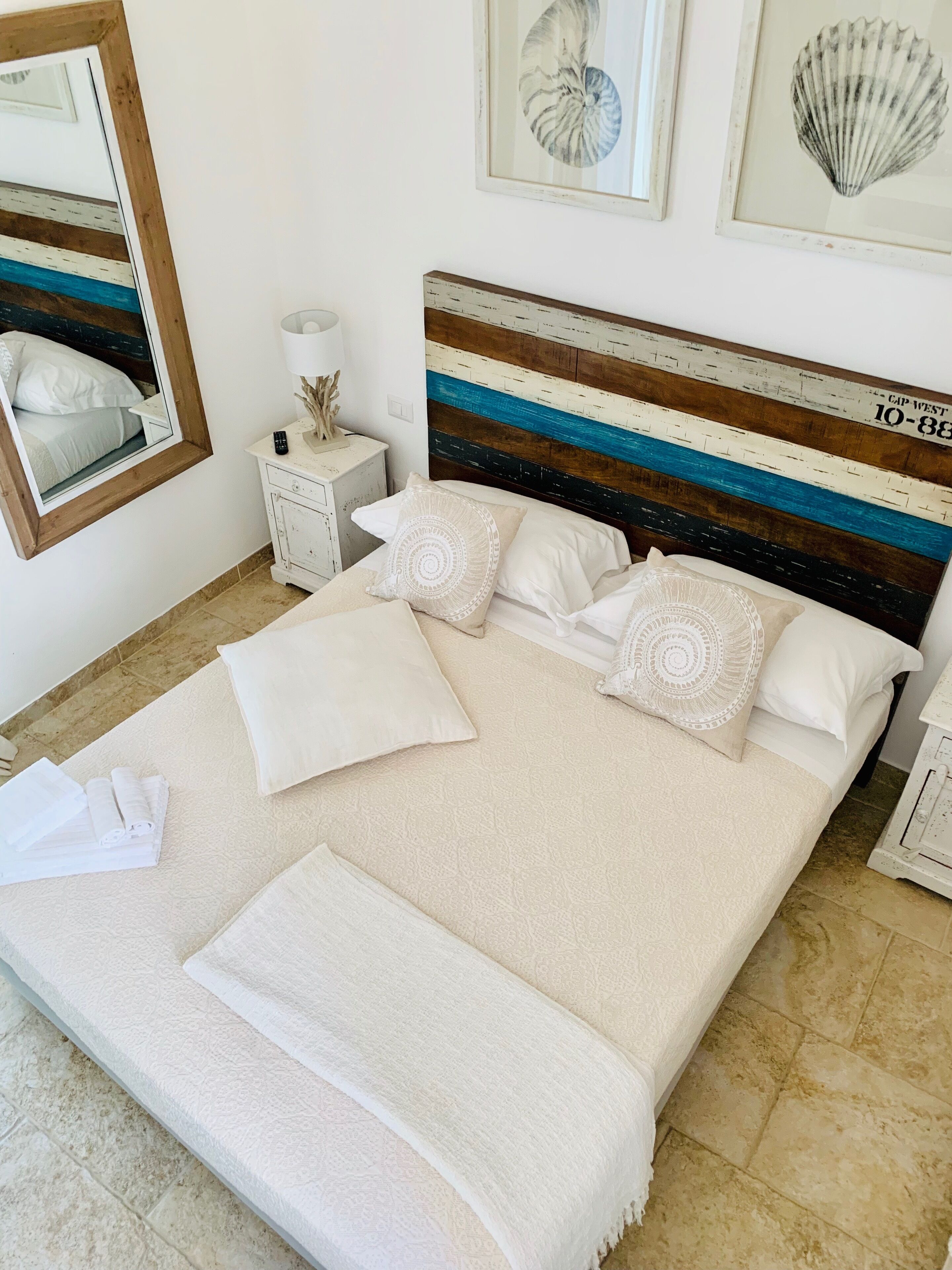 A neatly made bed with white linens and decorative pillows, a bedside table with a lamp and a small plant, and a wall with framed artwork.