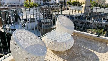 A rooftop terrace with two white, perforated chairs and a glass table, surrounded by a metal railing.