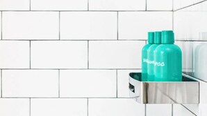 Une salle de bain avec des murs recouverts de carreaux blancs et un lavabo en chrome, sur lequel se trouve une bouteille de shampoing verte.