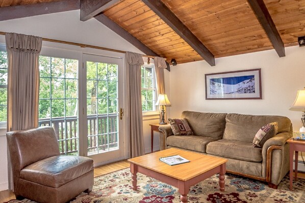 Una sala de estar acogedora con techo de madera, un sofá de cuero marrón, una mesa de centro de madera y un gran ventanal con cortinas.
