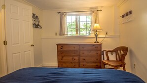Ein Schlafzimmer mit Bett, Kommode, Stuhl, Fenster mit Vorhängen und einer Lampe.