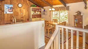 Ein rustikales Blockhaus mit einem Essbereich aus Holz, einer Küche mit einer Uhr und einem Schild mit der Aufschrift ‚Wildcat‘, sowie einer Treppe, die zu einem Schlafboden führt.