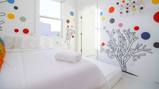 Une chambre moderne avec un grand lit, du linge de lit blanc, une fenêtre et des décorations murales colorées, notamment un arbre et des cercles.