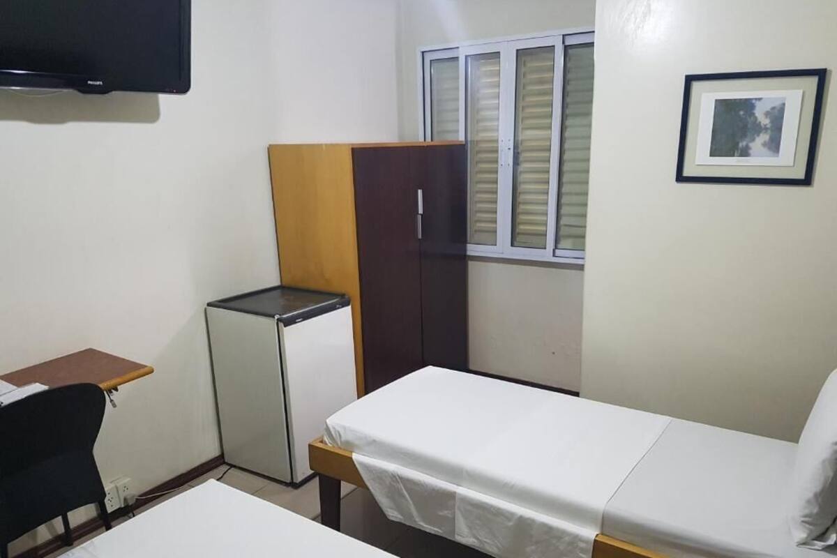A small, clean room with a bed, a desk, a wardrobe, and a window with blinds.