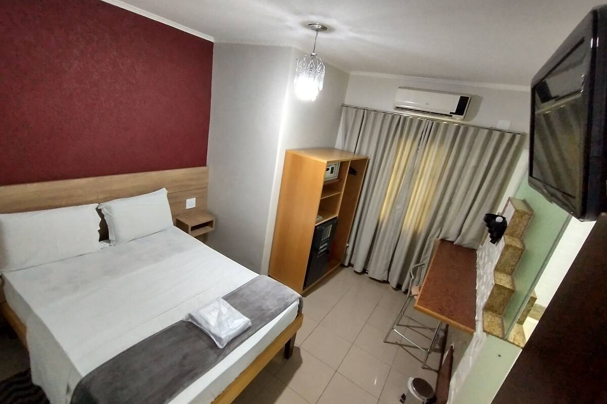 A hotel room with a bed, a wooden nightstand, a small wooden shelf, a desk with a chair, a television, and an air conditioning unit.