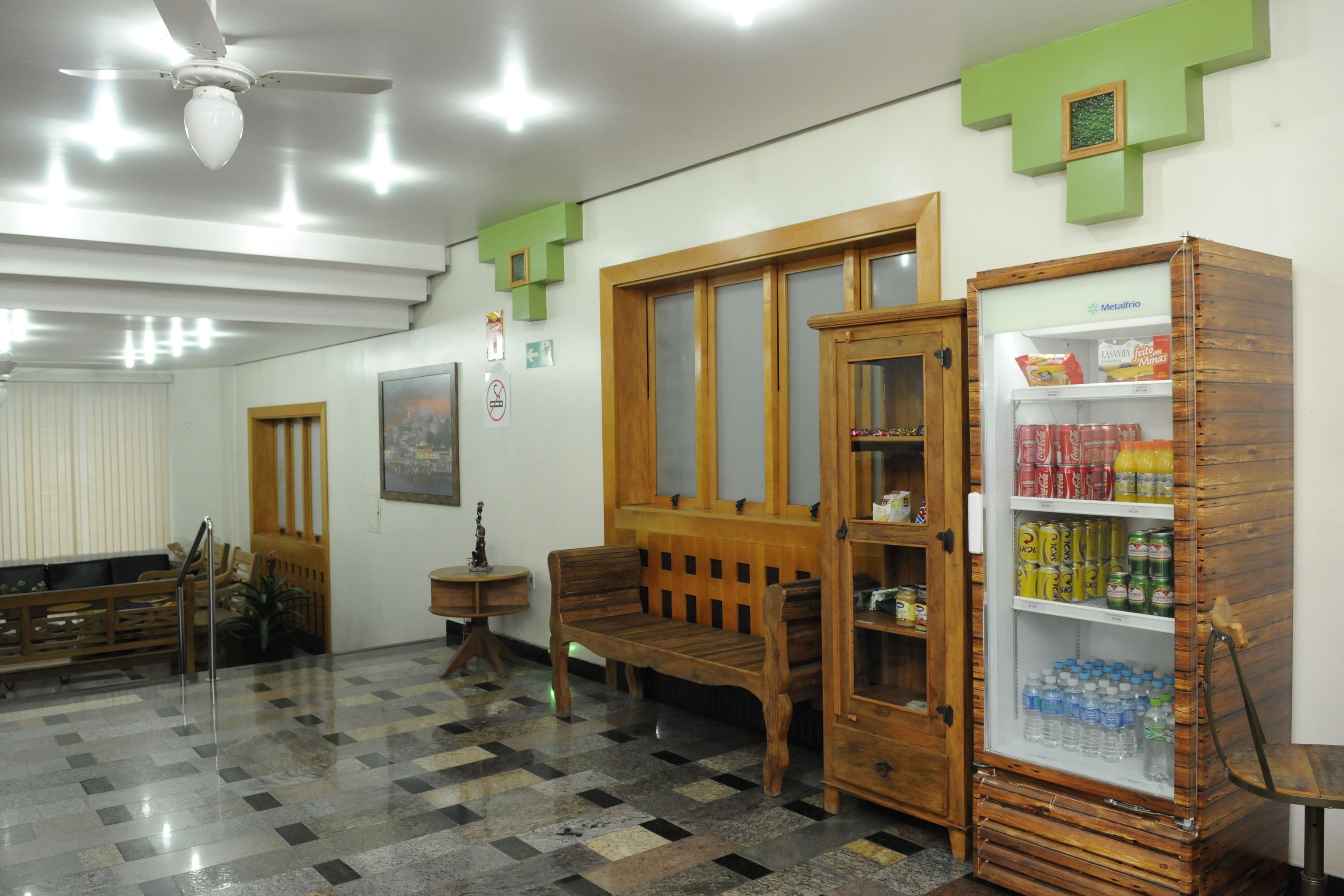 Um hall com piso xadrez, mobiliário de madeira, geladeira abastecida com bebidas e um ventilador de teto.
