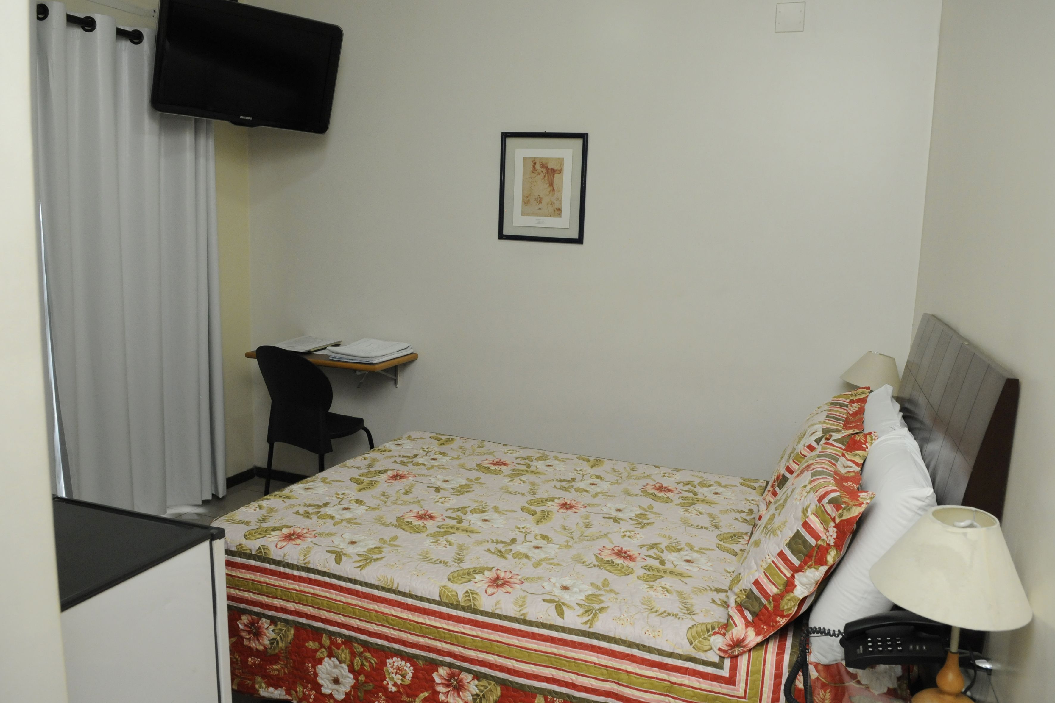 Una habitación de hotel con una cama, un escritorio con una silla, un televisor montado en la pared y una lámpara de noche.