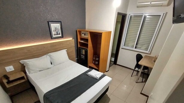 A hotel room with a bed, a desk, a chair, a wardrobe, and a framed picture on the wall. - Hotel Cidade (Passos)