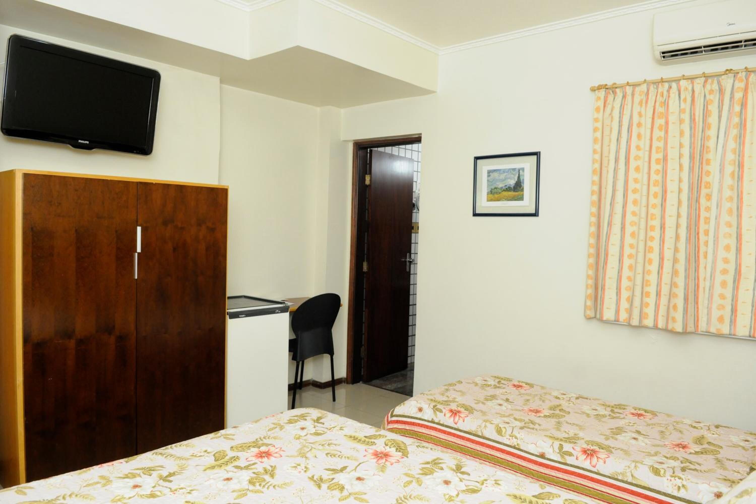 A room with a bed, a desk, a chair, a wardrobe, a television, and a window with curtains.
