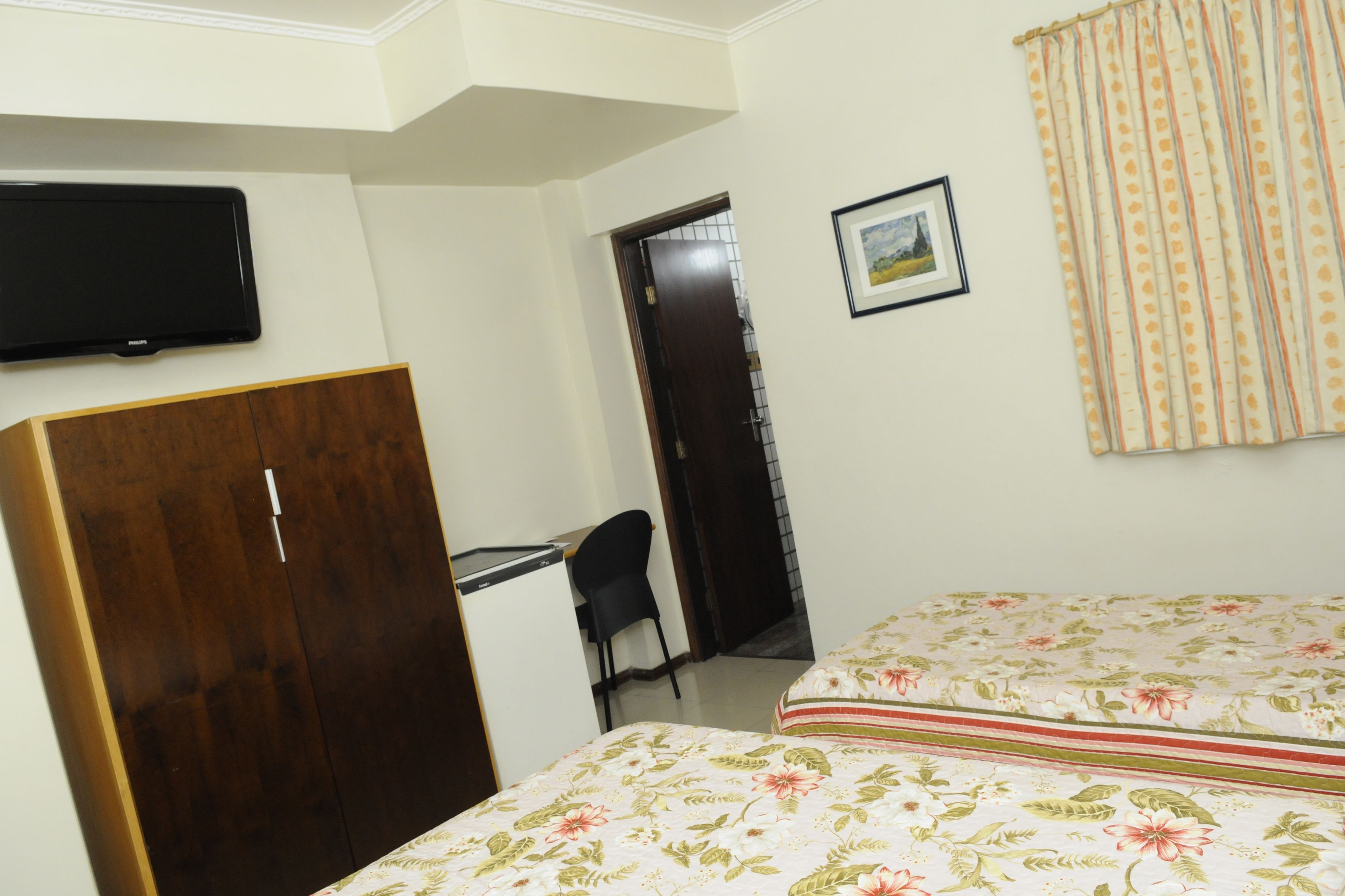 A bedroom with a bed, a desk, a chair, a wardrobe, a television, and a door leading to another room.