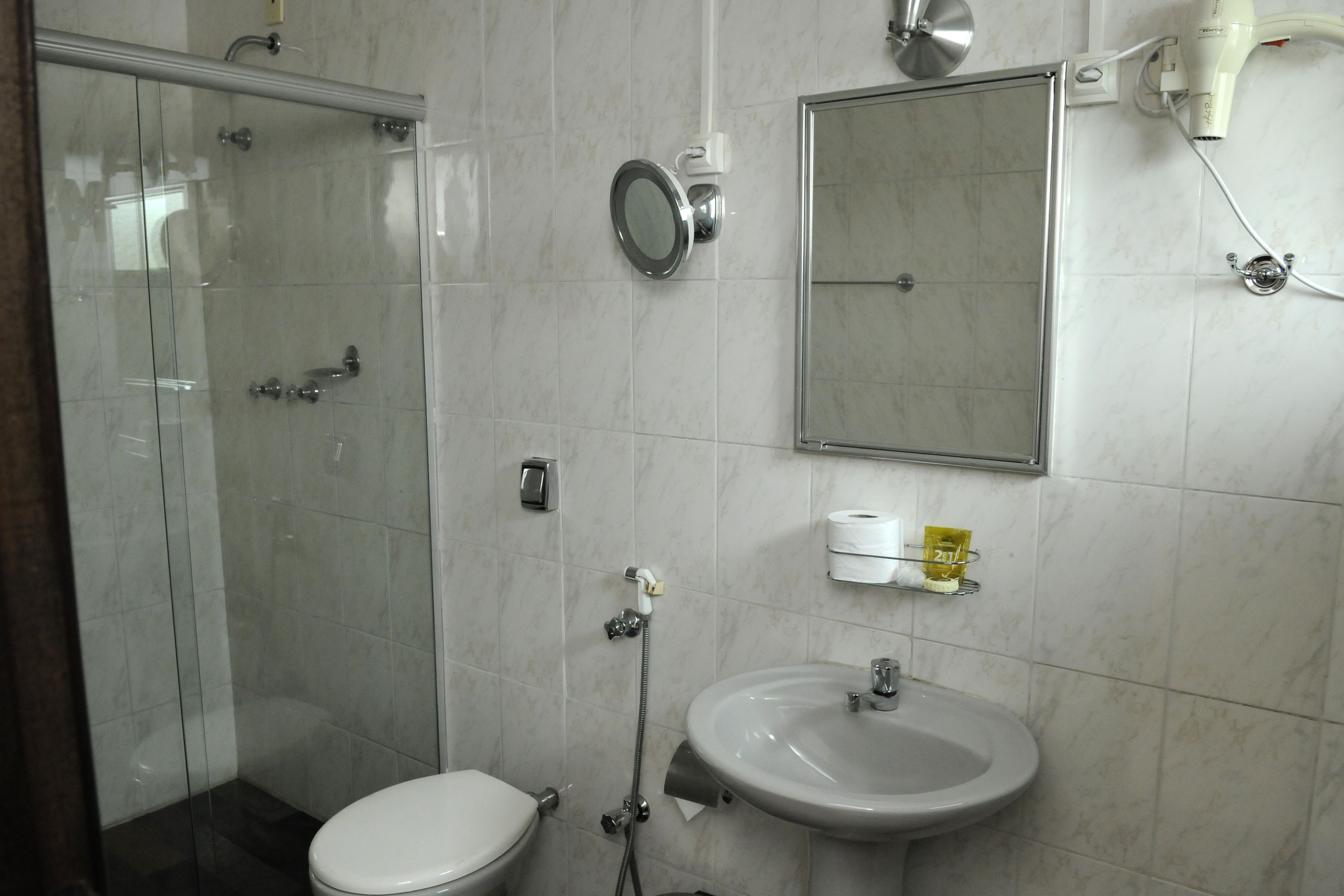 A bathroom with a glass shower enclosure, a toilet, a sink, a mirror, and a hairdryer on the wall.
