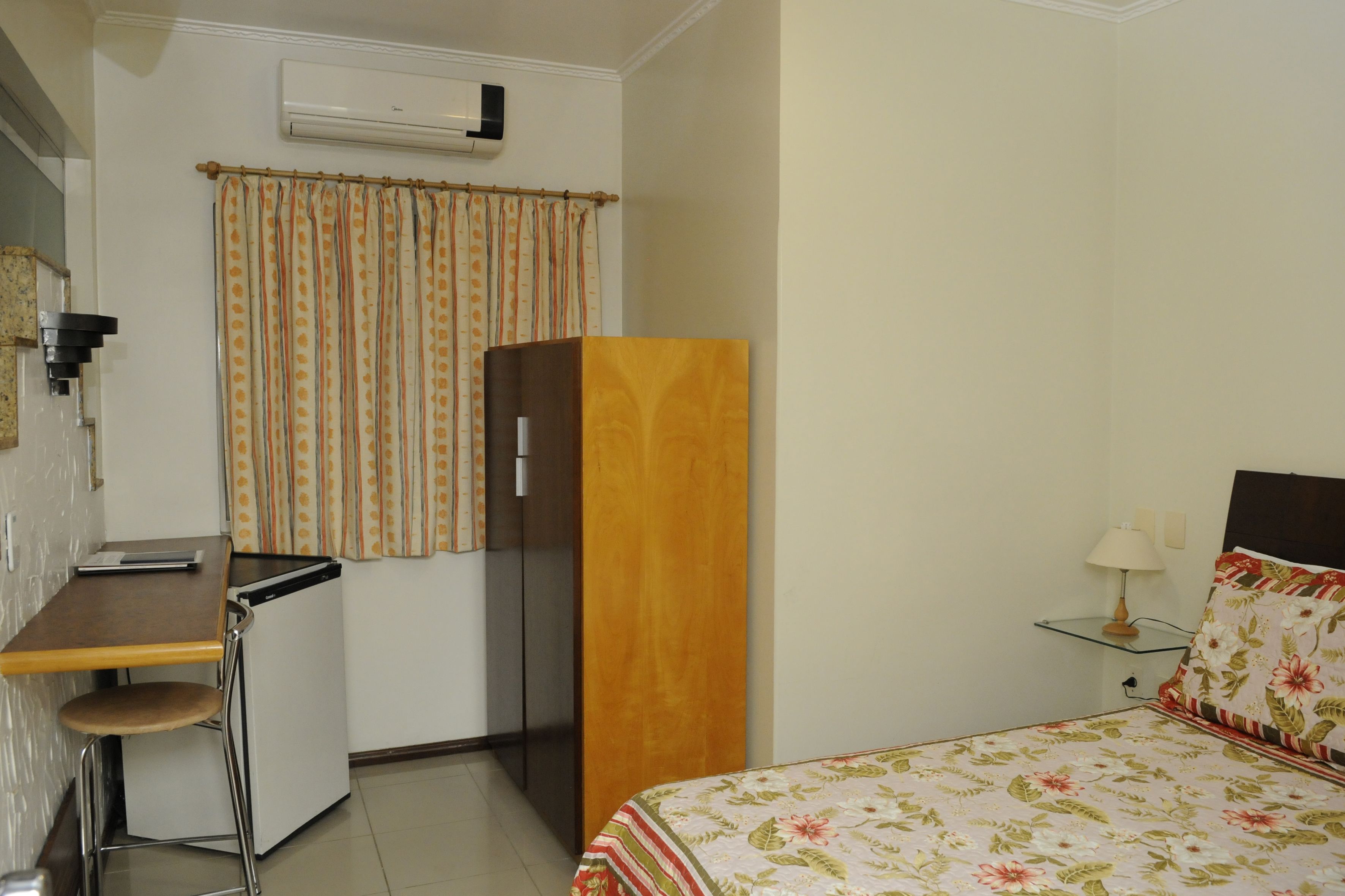 Habitación de hotel compacta con kitchenette, cama con ropa de cama floral, armario de madera y una mesita de noche pequeña con lámpara.