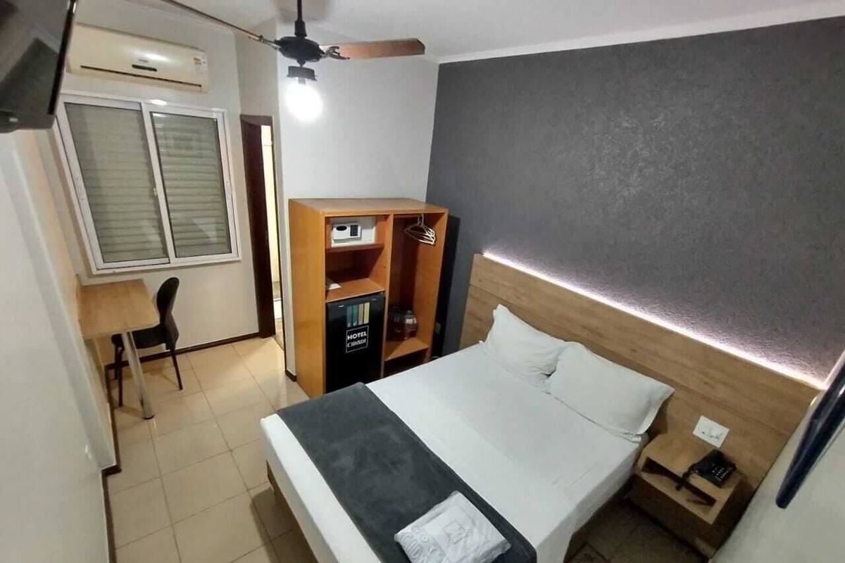 A hotel room with a bed, bedside table, nightstand, wardrobe, desk, chair, and an air conditioning unit.