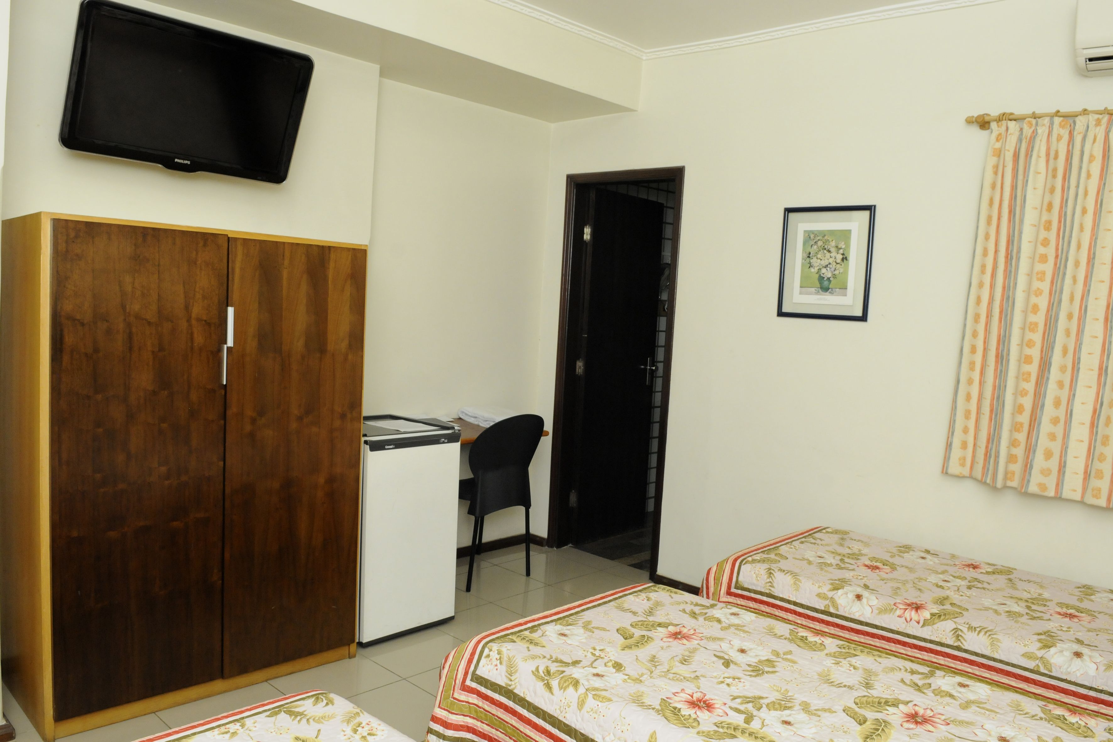 Ein Zimmer mit Holzschrank, Schreibtisch mit Stuhl, Bett mit Blumenmuster und Fernseher an der Wand, sowie einem gerahmten Bild.