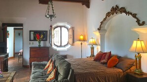 A cozy room with a bed, a sofa, and a wooden ceiling. There is a window with a view, a lamp, and a vase with flowers. - Casa Hermano Pedro (Antigua Guatemala)