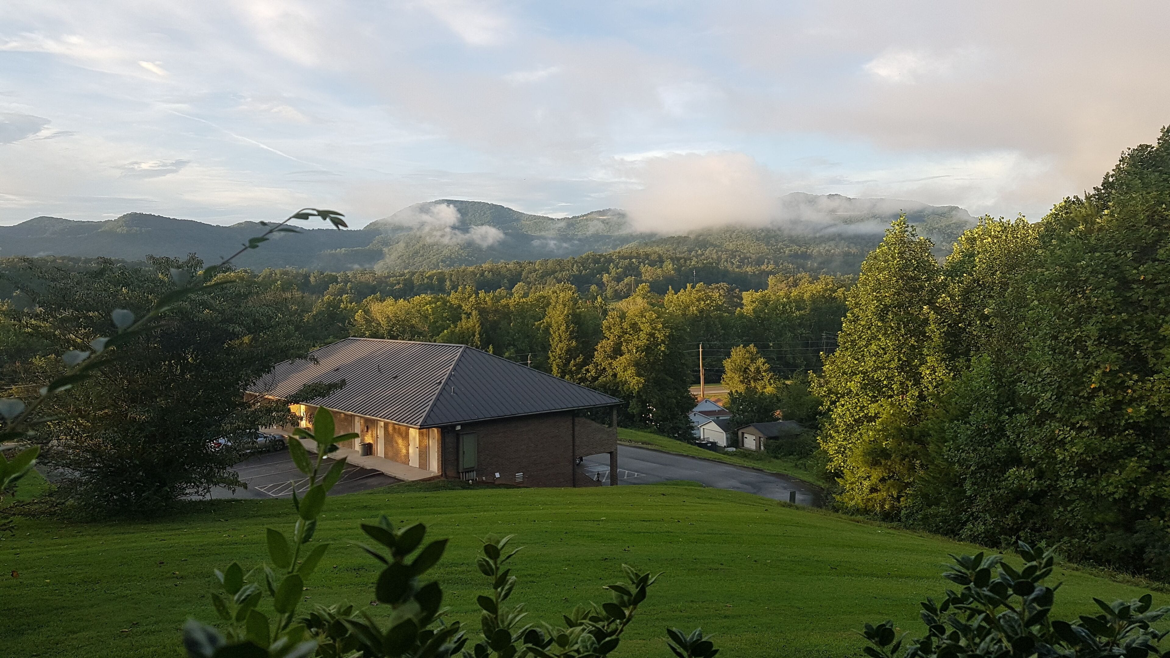 A house with a sloped roof and a brick exterior, surrounded by lush greenery and a hilly landscape.
