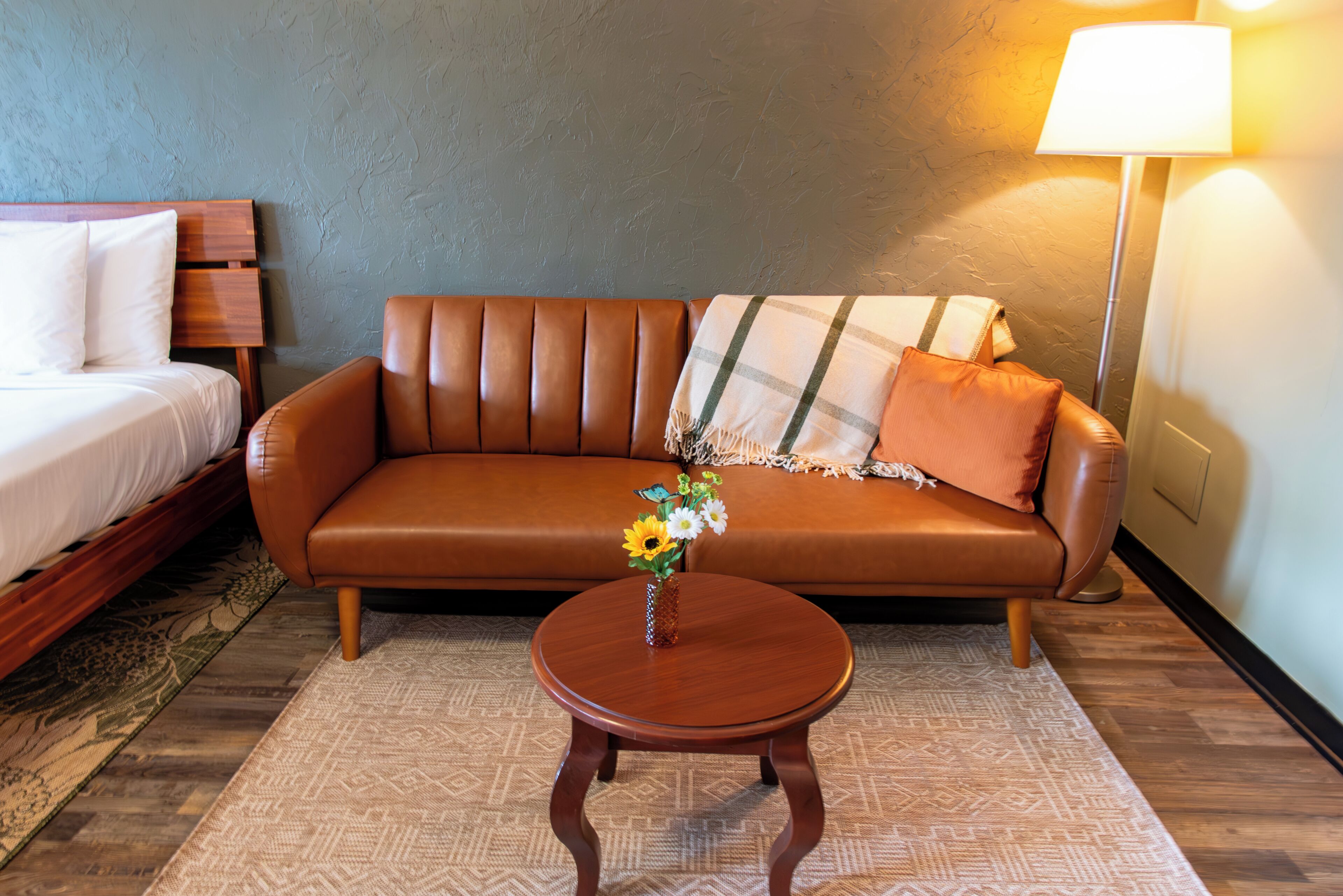 Una habitación con un sofá de cuero marrón, una cama de madera, una mesita de madera con un jarrón de flores y una lámpara.