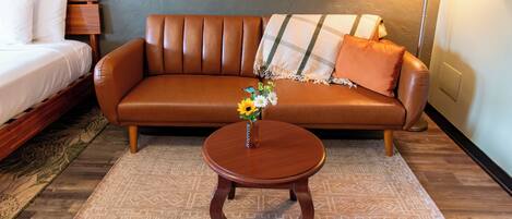 Una habitación con un sofá de cuero marrón, una cama de madera, una mesita de madera con un jarrón de flores y una lámpara.