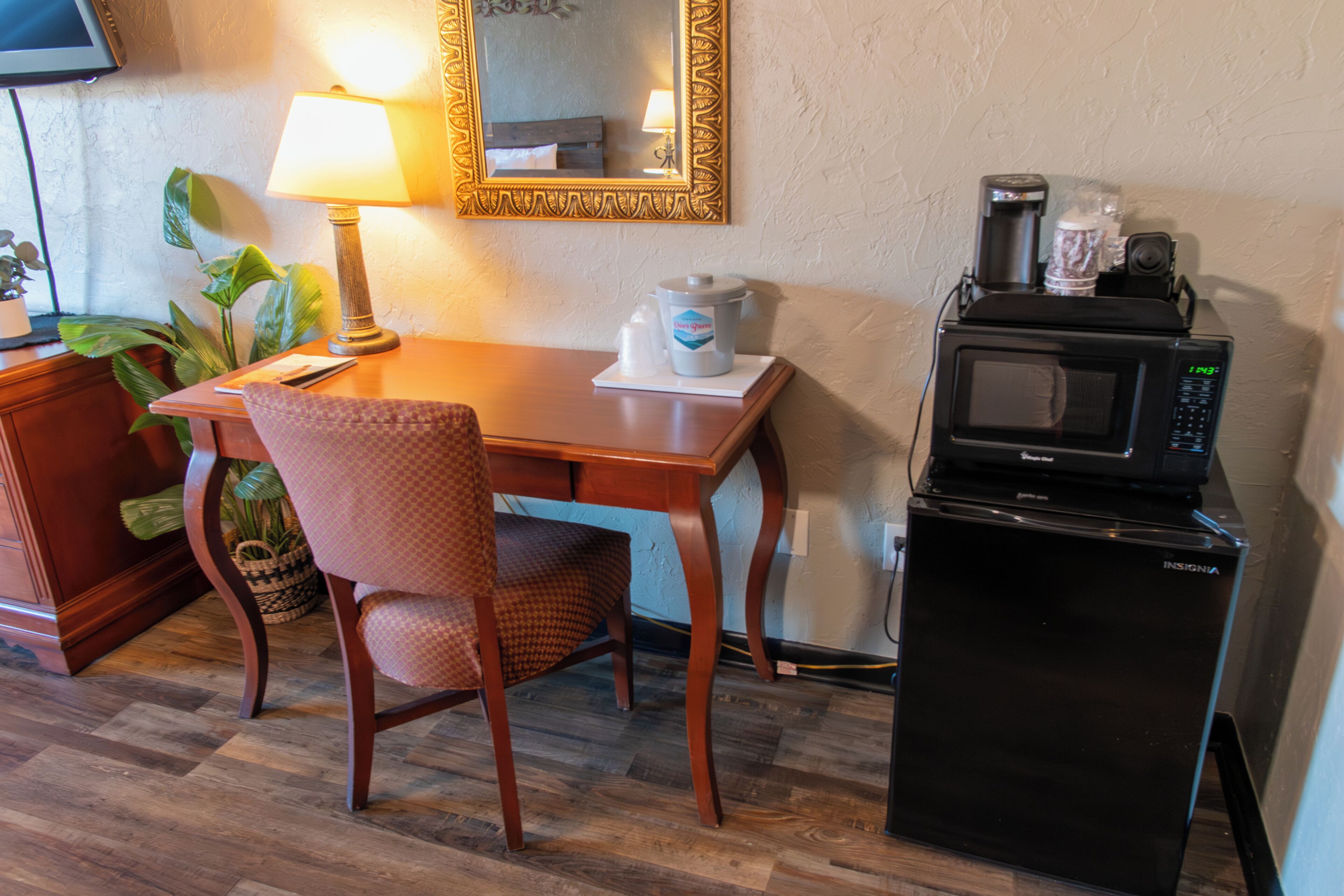 Kompakti hotellihuone, jossa on puinen työpöytä, tuoli, pieni jääkaappi, mikroaaltouuni, kahvinkeitin ja lamppu.