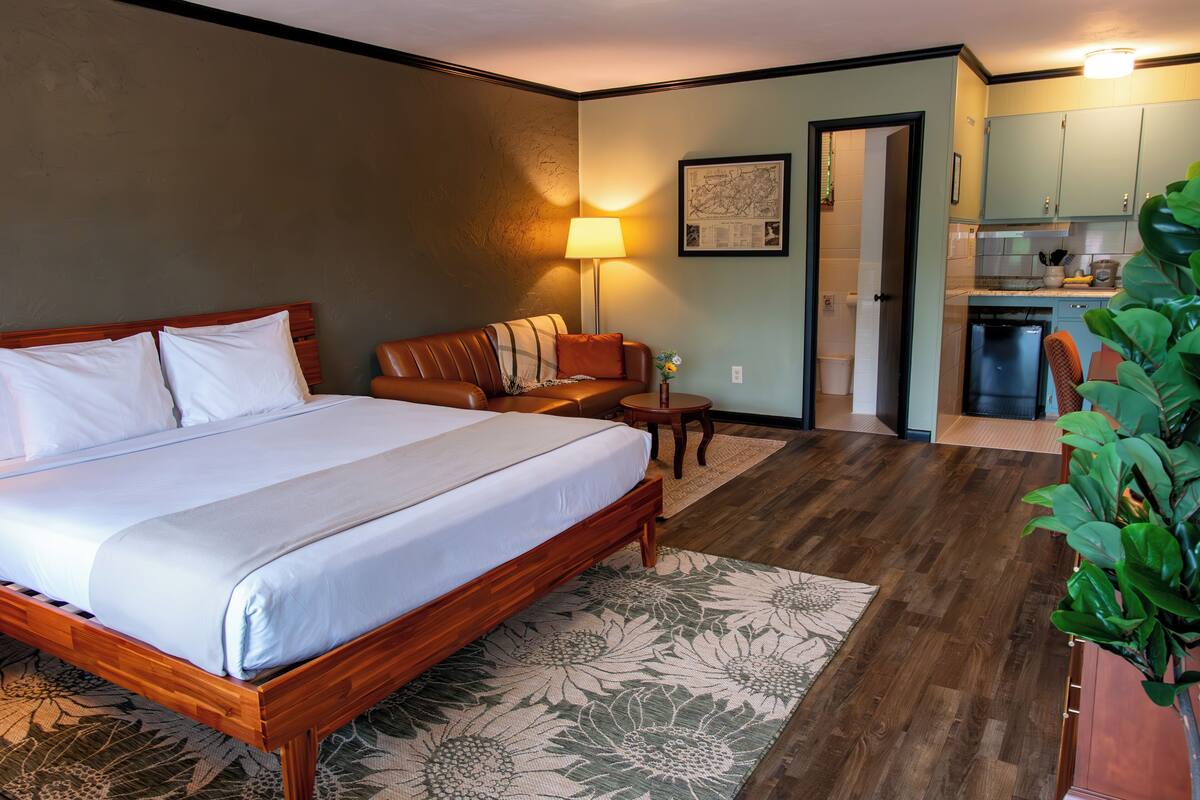 A hotel room with a large bed, a sofa, a small table, a lamp, and a kitchen area visible through an open door.