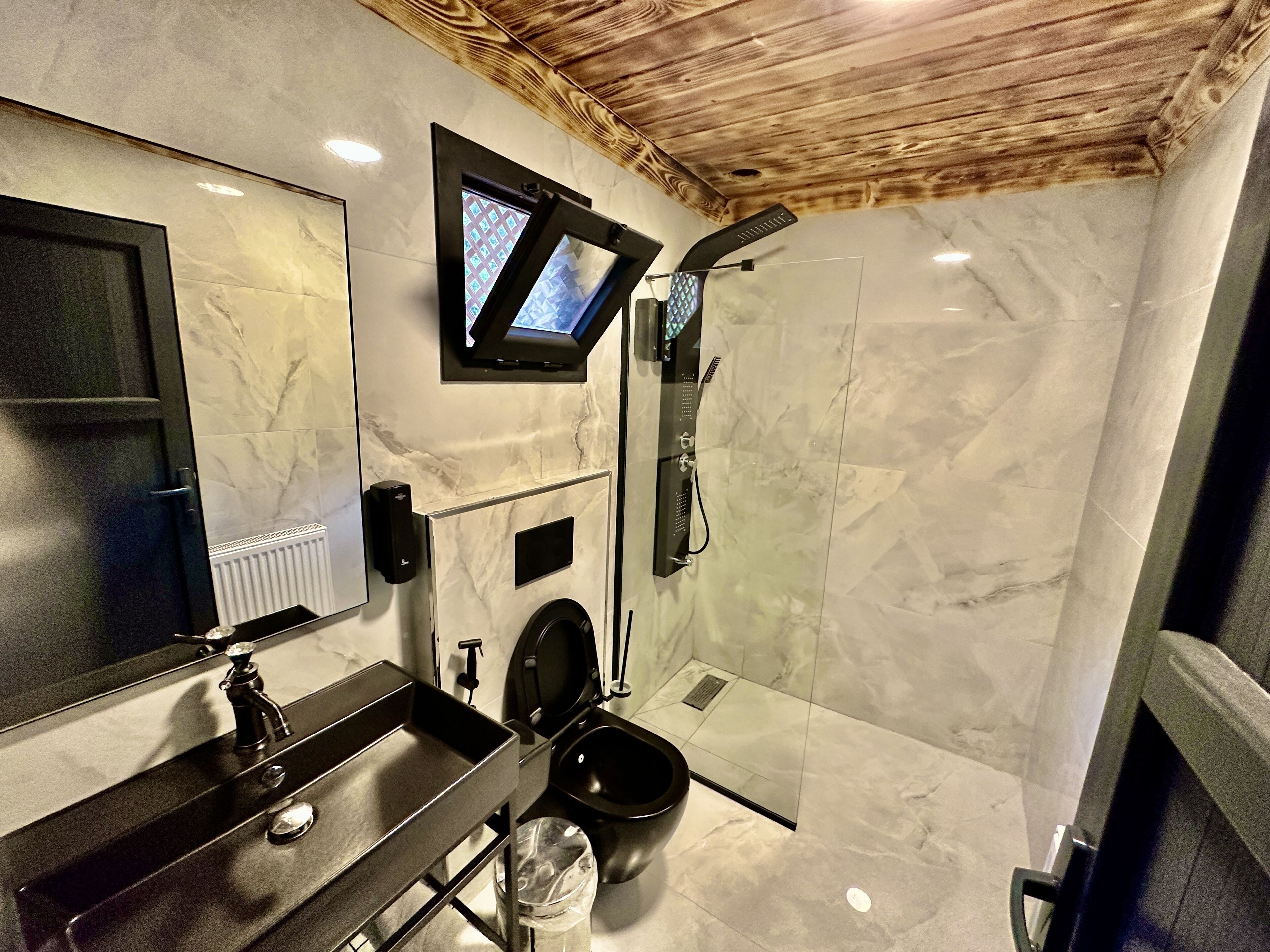 A modern bathroom with a marble floor, a black sink, a toilet, a shower with a handheld showerhead, and a television mounted on the wall.