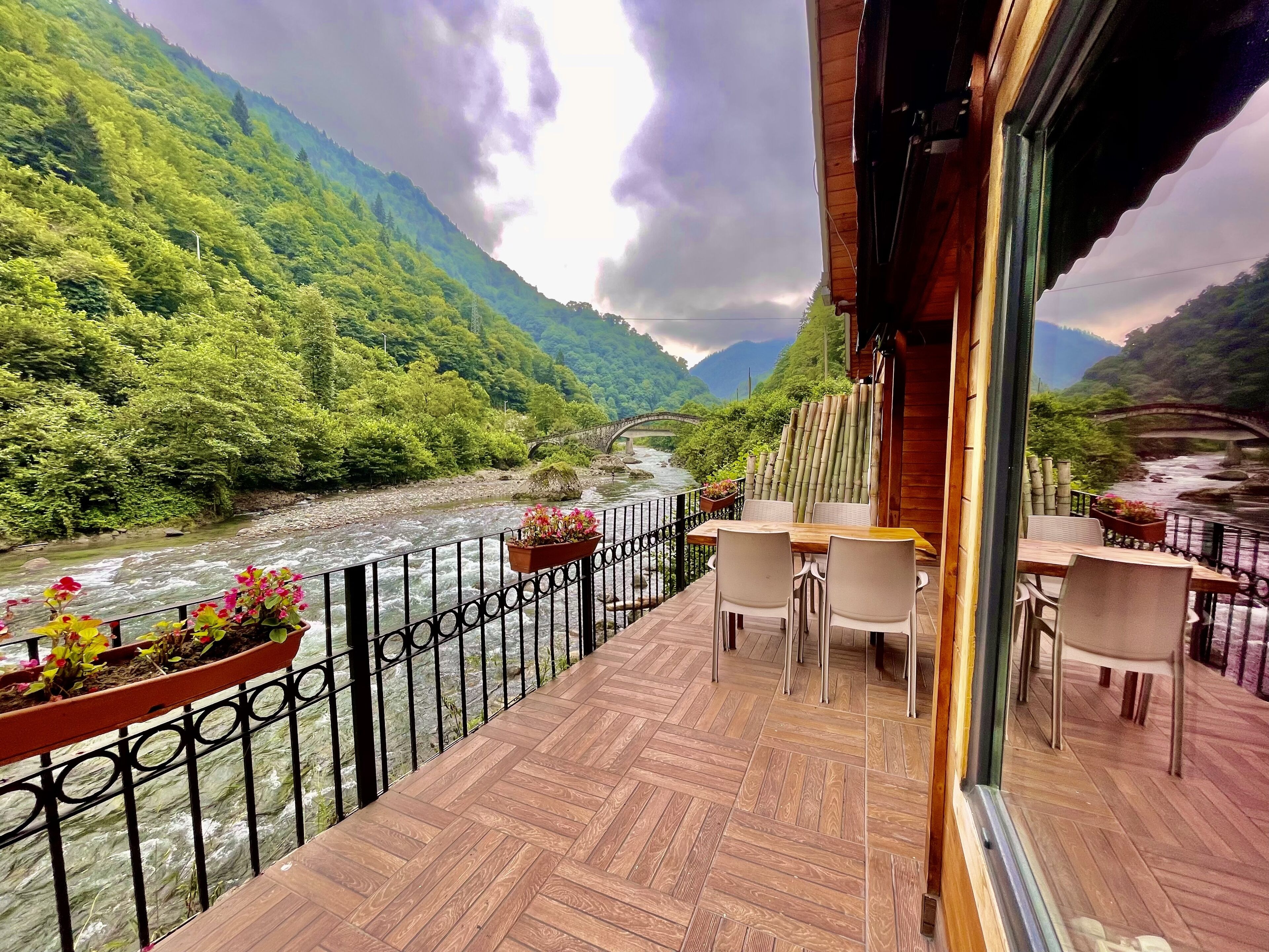 Un balcon doté d’un plancher en bois, de chaises blanches et d’une table, offrant une vue sur une rivière et un pont.