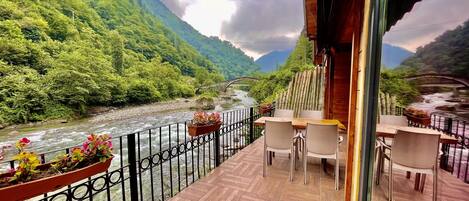 A balcony with wooden flooring, white chairs, and a table overlooking a river with a bridge.