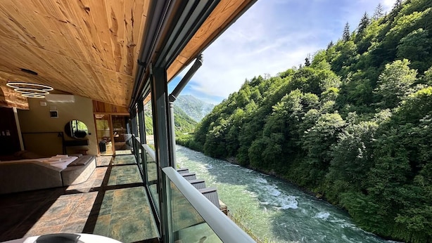 Habitación con pared de vidrio con vistas a un río, techo de madera y zona de estar con sofá y mesa.