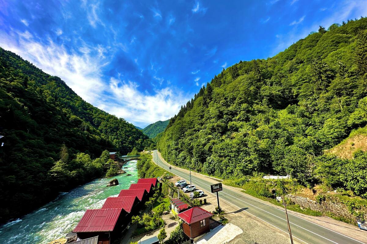 Un hôtel situé au bord de la rivière, avec des bâtiments aux toits rouges, une route qui passe à proximité et, en arrière-plan, de luxuriantes collines verdoyantes.