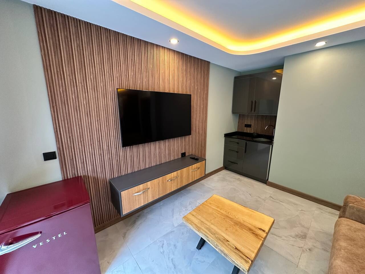 Ein modernes Wohnzimmer mit einem an einer Holzvertäfelung befestigten Flachbildfernseher, einem Holztisch und einer kleinen Küchenzeile im Hintergrund.