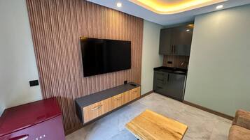 Ein modernes Wohnzimmer mit einem an einer Holzvertäfelung befestigten Flachbildfernseher, einem Holztisch und einer kleinen Küchenzeile im Hintergrund.
