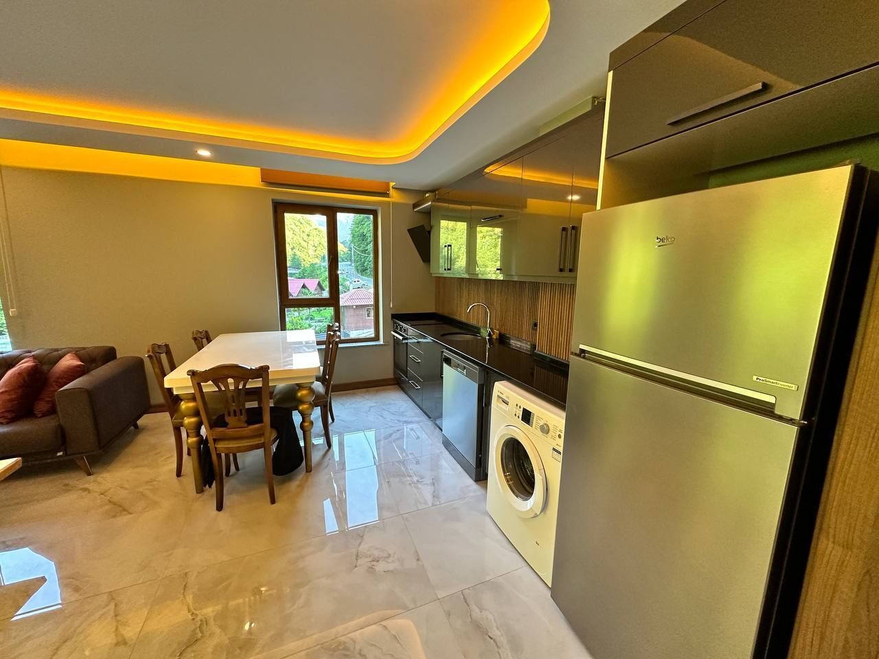 A modern kitchen with a stainless steel refrigerator, a built-in washing machine, and a dining area with a table and chairs.
