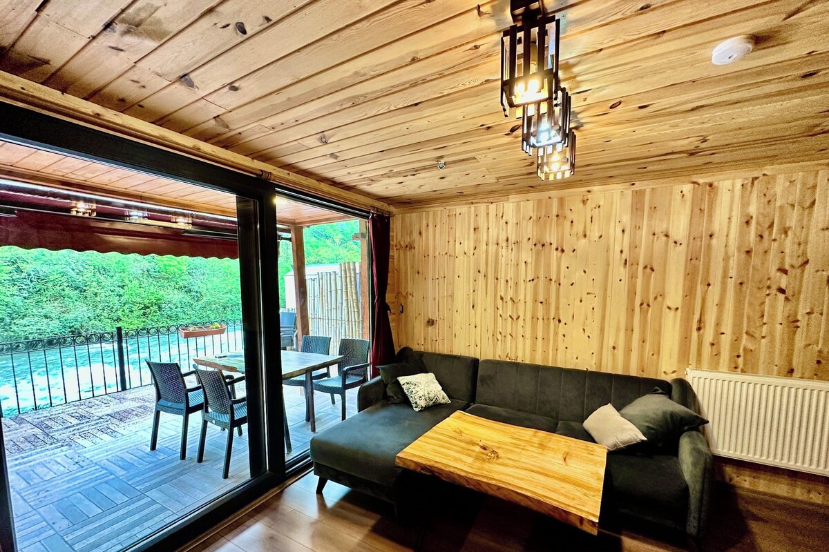 Una habitación acogedora con techo de madera, un sofá, una mesa y vistas a un jardín a través de amplias ventanas.