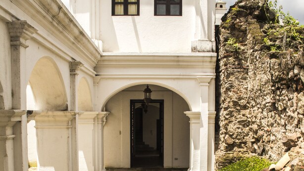 A white building with a cobblestone pathway leading to a dark entrance, surrounded by stone walls and greenery. - Casa Las Ruinas (Antigua Guatemala)