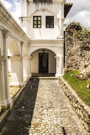 Un edificio blanco con un sendero adoquinado que lleva a una entrada oscura, rodeado de muros de piedra y follaje.