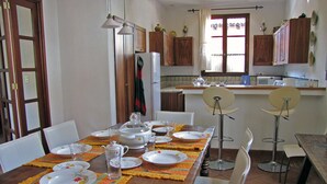 A dining area with a wooden table set for four, surrounded by white chairs. There are two bar stools, a refrigerator, and wooden cabinets in the background. - Casa Real (Antigua Guatemala)