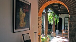 A room with a desk, a framed artwork, and a brick archway leading to an outdoor area with plants and a bench. - Casa Real (Antigua Guatemala)