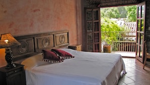 A bedroom with a large bed, a nightstand with a lamp, a wooden headboard, and a view of a balcony with plants. - Casa Real (Antigua Guatemala)