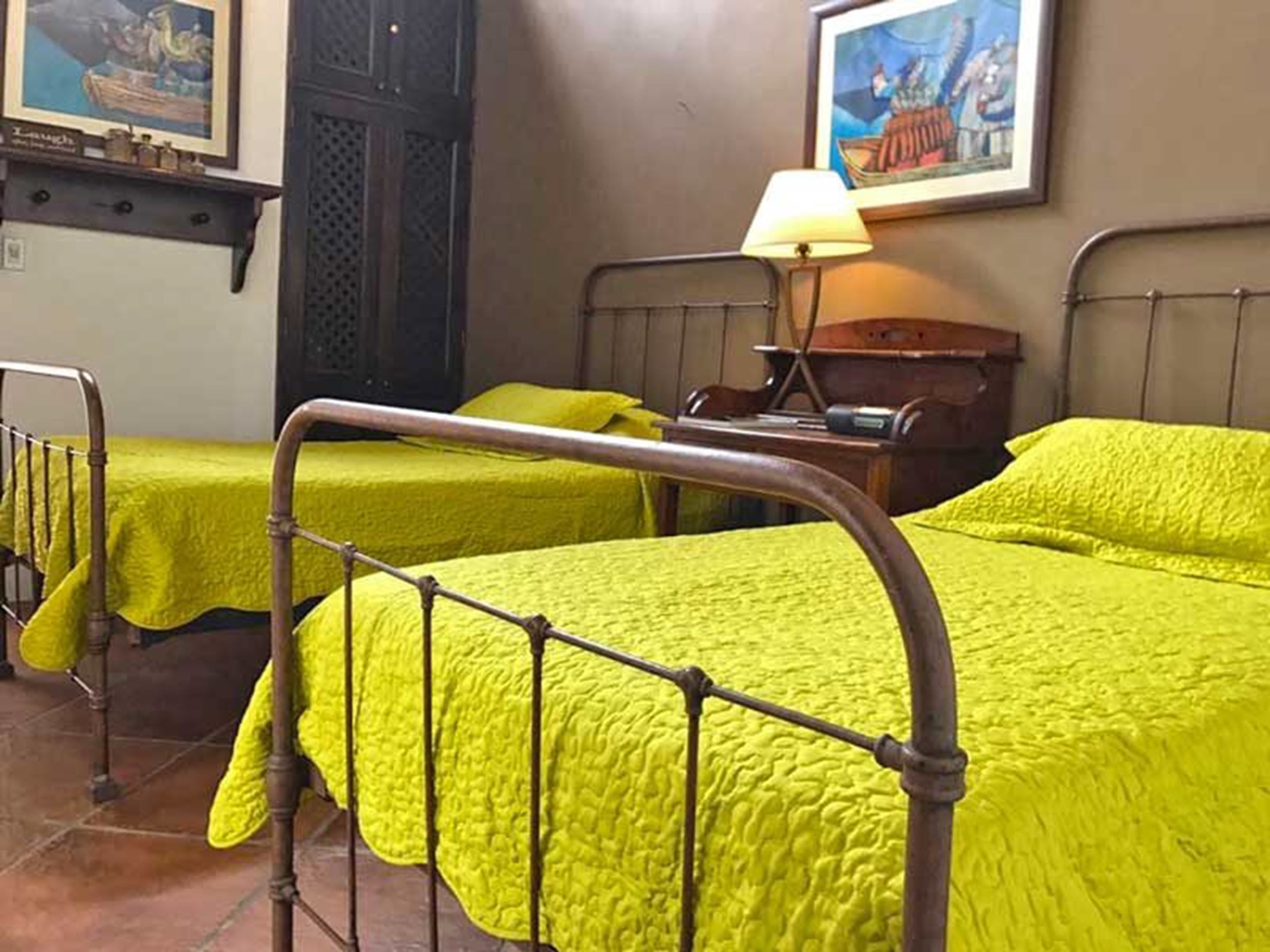 A room with two beds, each with a metal frame and bright green bedding. There is a lamp on a wooden nightstand beside one bed.