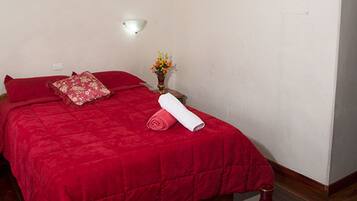 Een slaapkamer met een rood sprei, een klein tafeltje met een beeldje, een vaas en een plafondlamp.