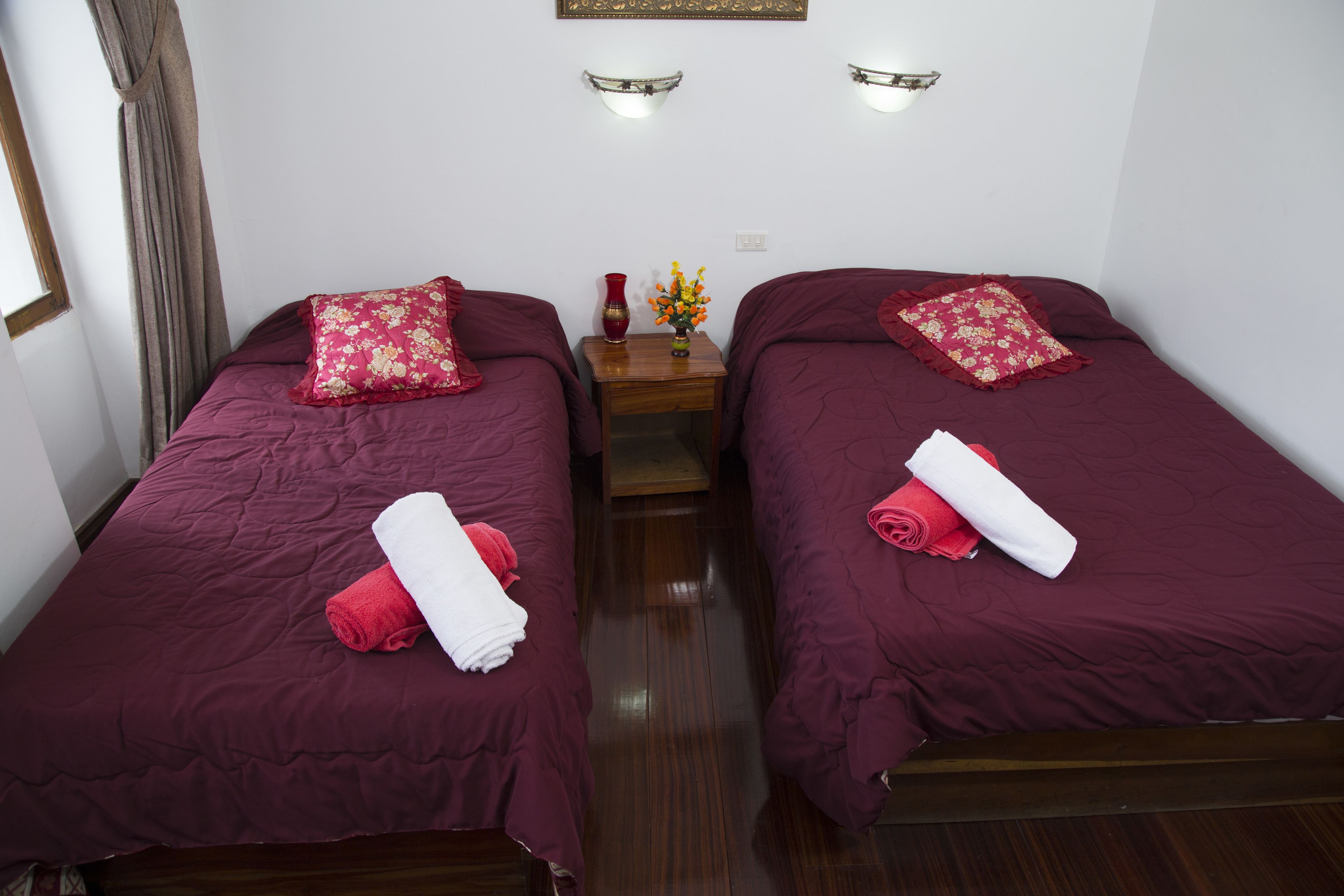 A hotel room with two single beds, each with a red bedspread and matching pillows. There is a wooden nightstand between the beds with a vase of flowers and a red candle.