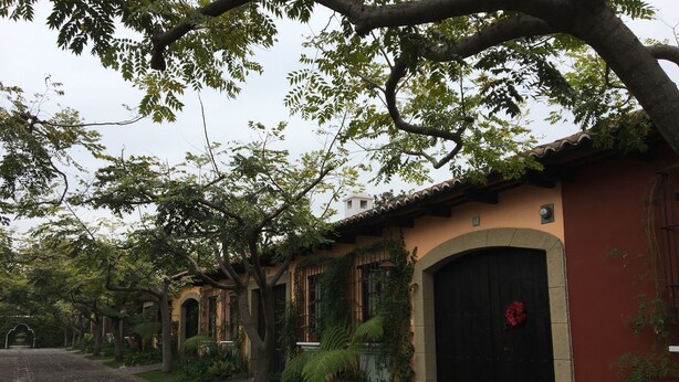 A cobblestone pathway leading to a row of traditional buildings with arched doorways and windows, surrounded by lush greenery. - Casa Del Calvario (Antigua Guatemala)